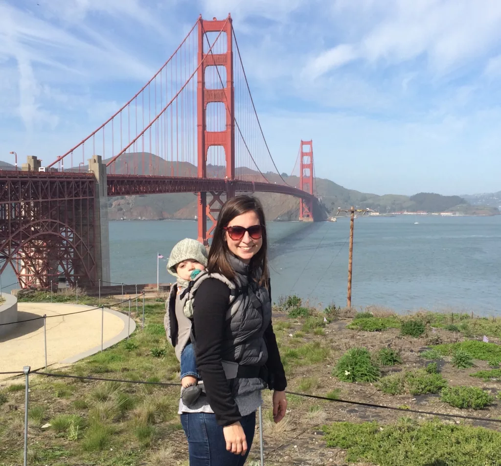 A woman standing in front of the Golden Gate Bridge with a baby in a baby carrier on her back.